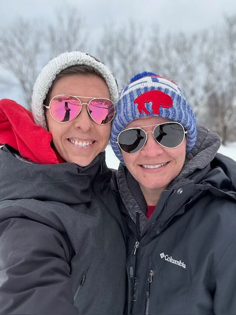 Two wives in beanie caps, black jackets, and aviator glasses smile in front of the snow-covered woods. Two wives in beanie caps, black jackets, and aviator glasses smile in front of the snow-covered woods.