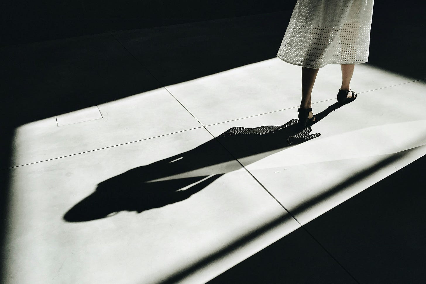 A woman in high heels and a white dress casts a long shadow. 