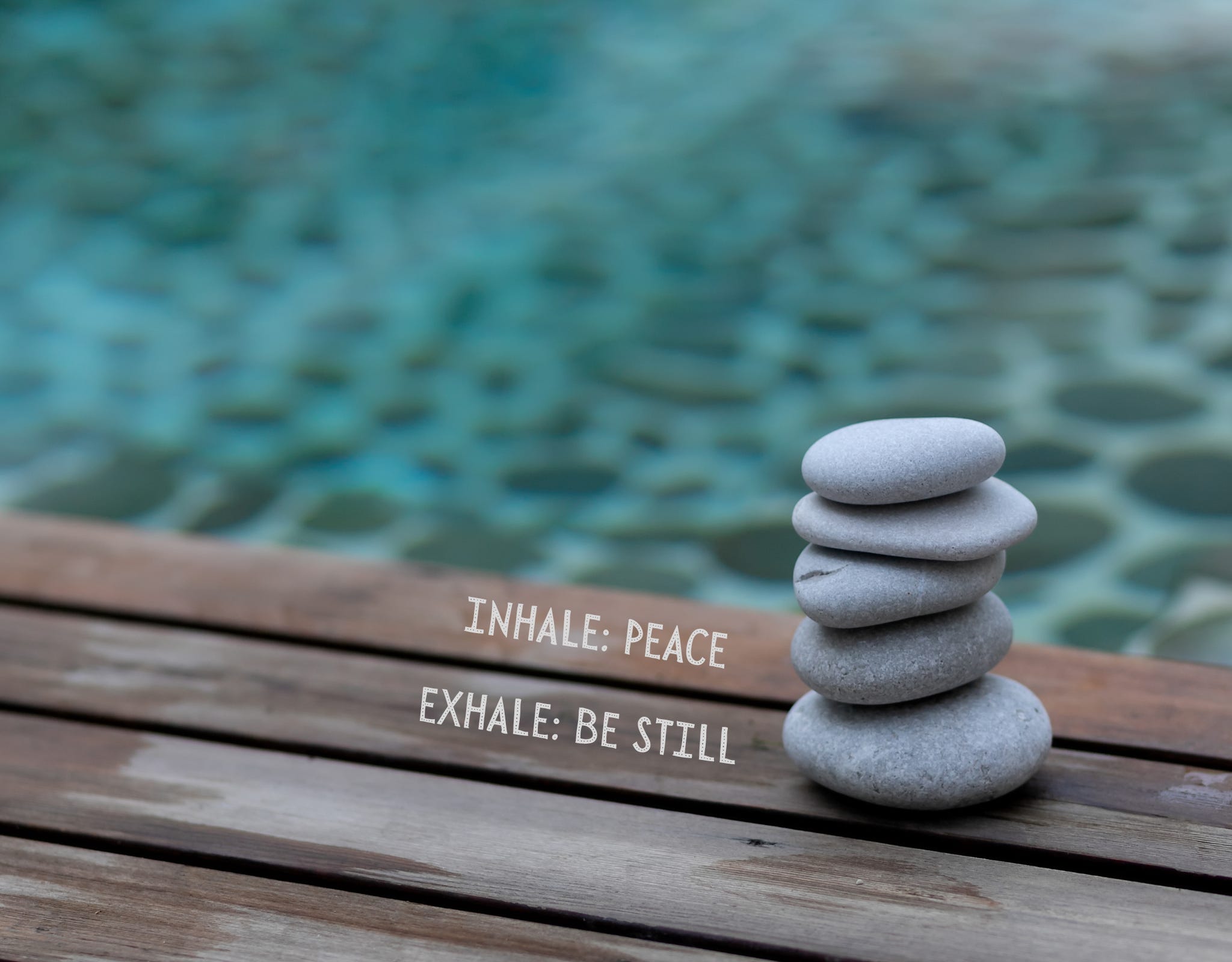 grey stones piled on top of each other sitting on a deck next to a pool. Words inscribed next to stones: Inhale: Peace. Exhale: Be Still