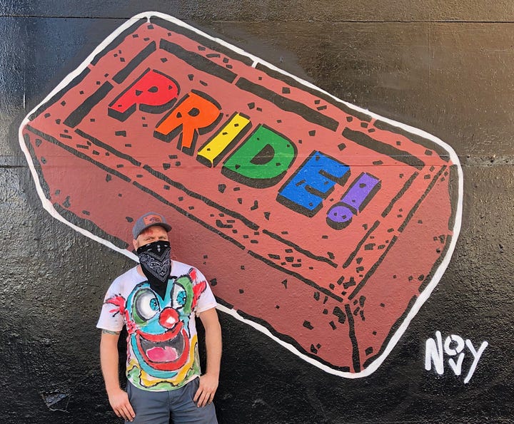 1) Jeremy is wearing a colorful shirt and bandana standing in front of a vibrant graffiti mural of a red brick with "PRIDE!" in rainbow letters, on top of a black wall, evoking a sense of celebration and diversity. Jeremy's signature and tag, Novy, is in white in the bottom right corner. Jeremy is wearing a white T-shirt with a clown face on it, and blue jeans. He is wearing a blue baseball cap and a black bandana over his nose and mouth. He has his thumbs in his pants pockets. 2) Colorful urban street art on a concrete structure, featuring a blue curled pattern, koi fish, and abstract designs. Nearby trees and parking sign visible. Jeremy's koi are on the far right pillar, and his signature and tag are on the bottom right of the pillar. It is attached to a white. building with several windows.