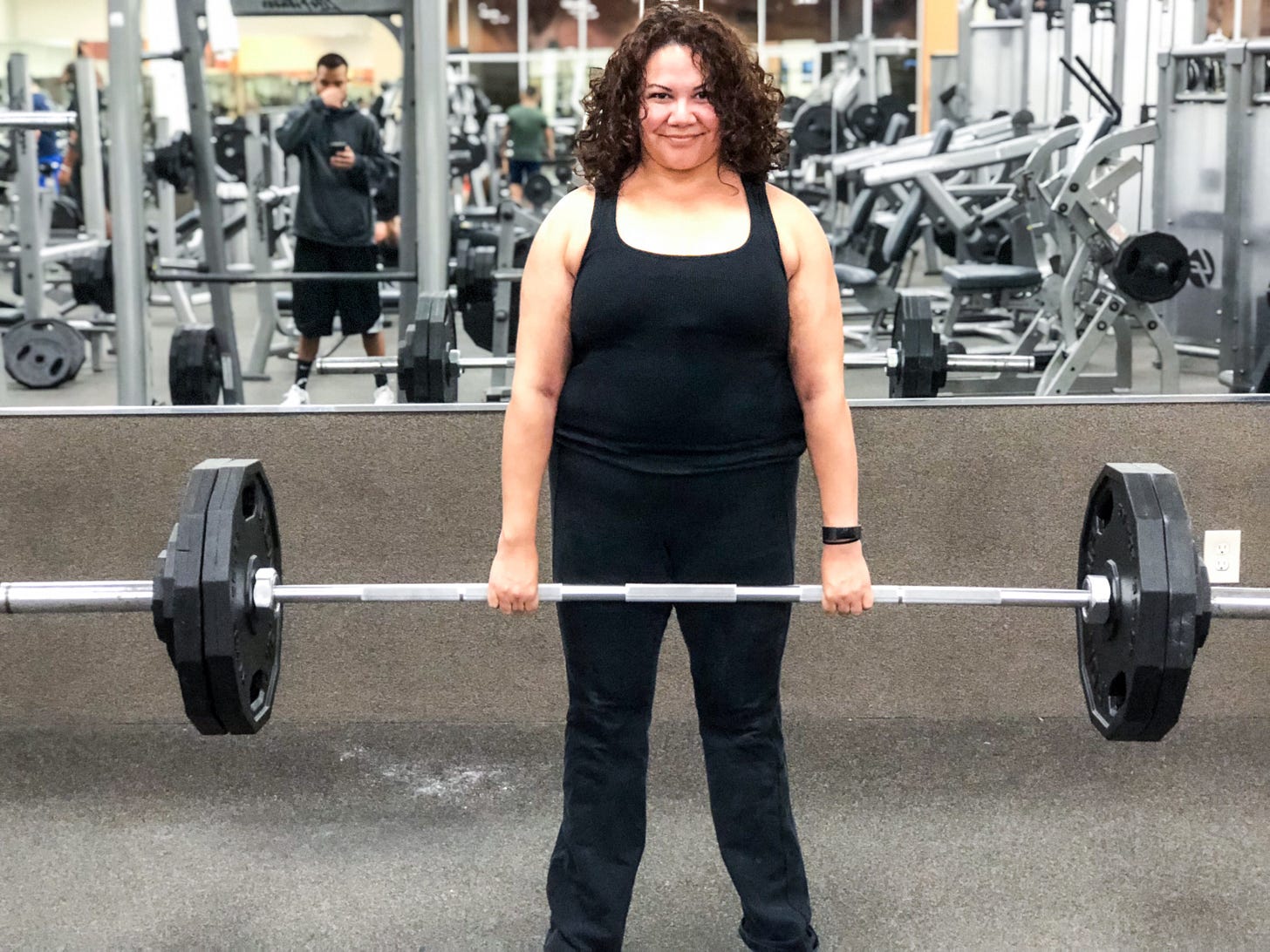 Paulette dressed in a black tank and black yoga pants is standing in the gym holding a barbell weighing 250 pounds in her bare hands