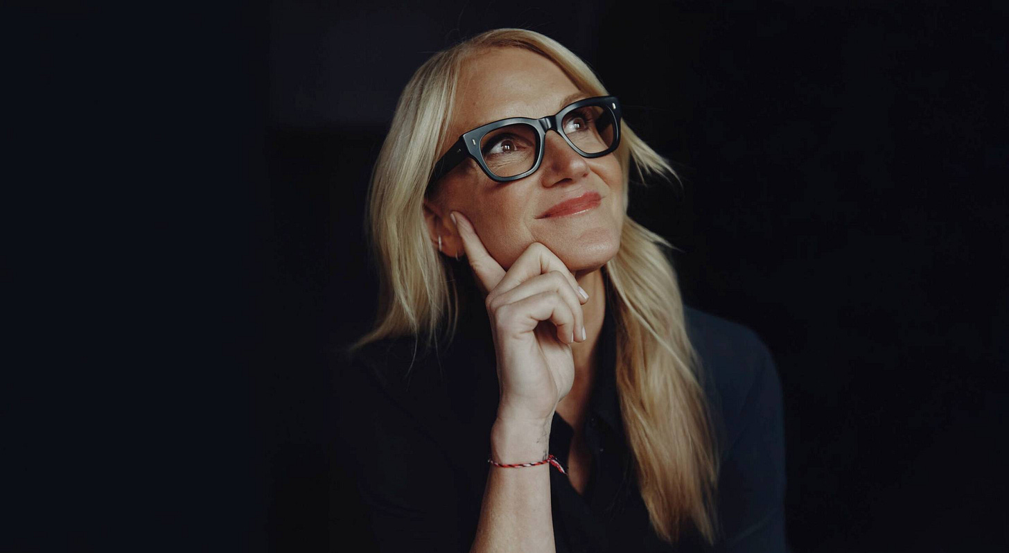 A woman with long blonde hair and glasses, resting her chin on her hand, appears thoughtful against a dark background.