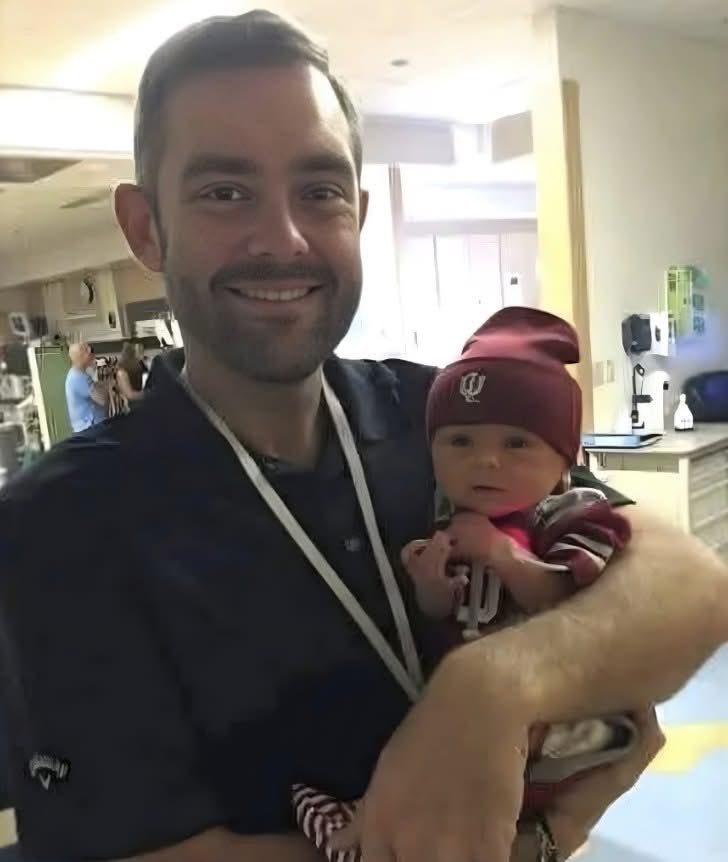 A man in a black polo shirt with a lanyard stands in a hospital room holding a newborn baby boy wrapped in a red and white blanket. The baby wears a red beanie and has a red Sooners outfit visible. Medical equipment and monitors are in the background, with another person blurred nearby. The setting includes a crib and window with blinds.