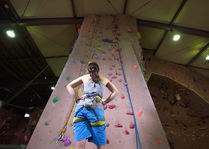 A woman standing at the base of a tall indoor rock wall looking up toward the ceiling