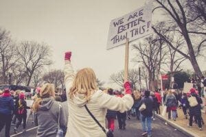 Photo of people protesting. A woman carries a sign reading, We are better than this!