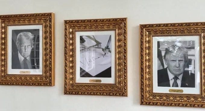 Three framed portraits of Donald Trump on a wall. Each portrait is in an ornate gold frame. The left and right portraits show Donald Trump in a suit, while the center portrait displays a document with a signature. Small plaques with text are visible below each portrait.