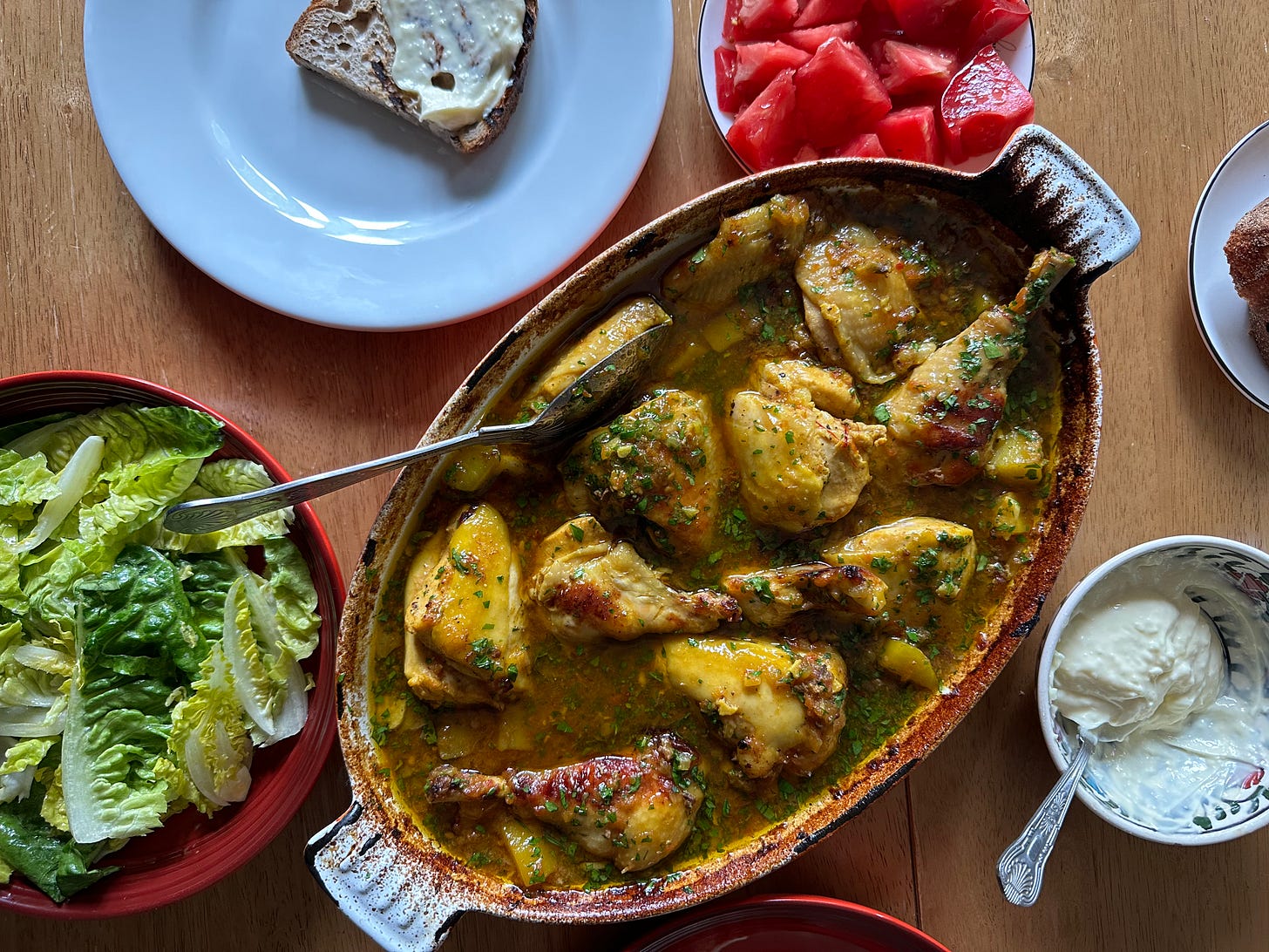 a table with a baking dish of Pastis and Saffron Chicken, bowls of lettuce, tomatoes, and aïoli, and a plate with a piece of bread and aïoli