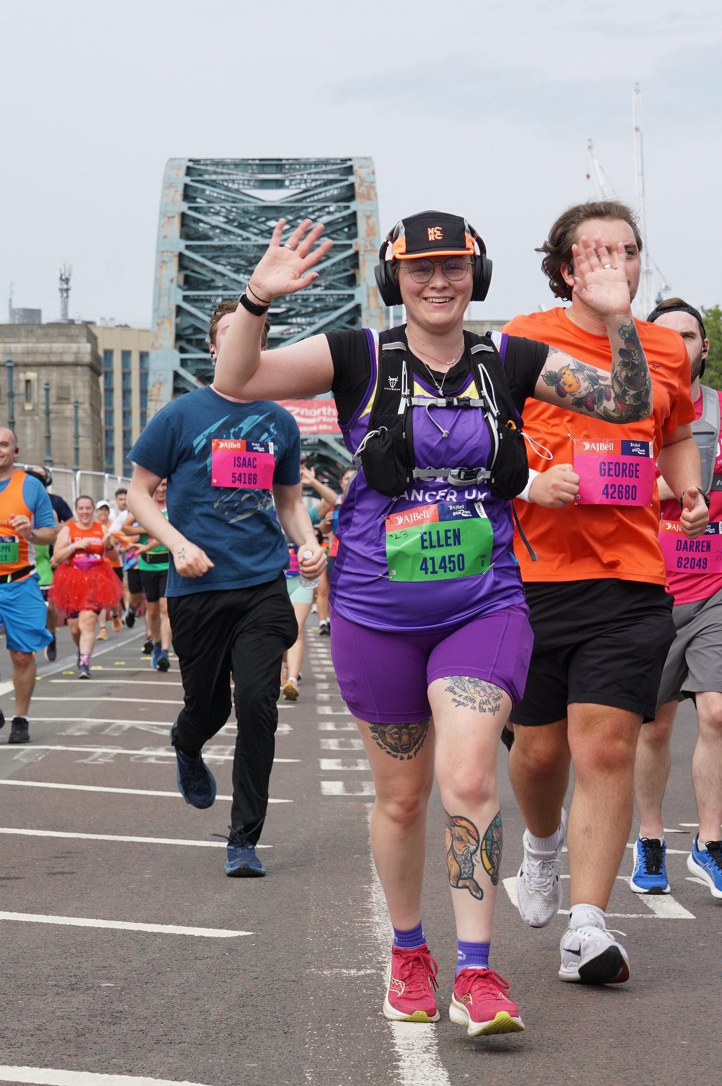 Event photo of Ellen with two arms in the air, waving at the photographer - she is wearing purple running gear, as a number pinned to her front, and is smiling. She is surrounded by other runners. In the background is Newcastle's Tyne Bridge.