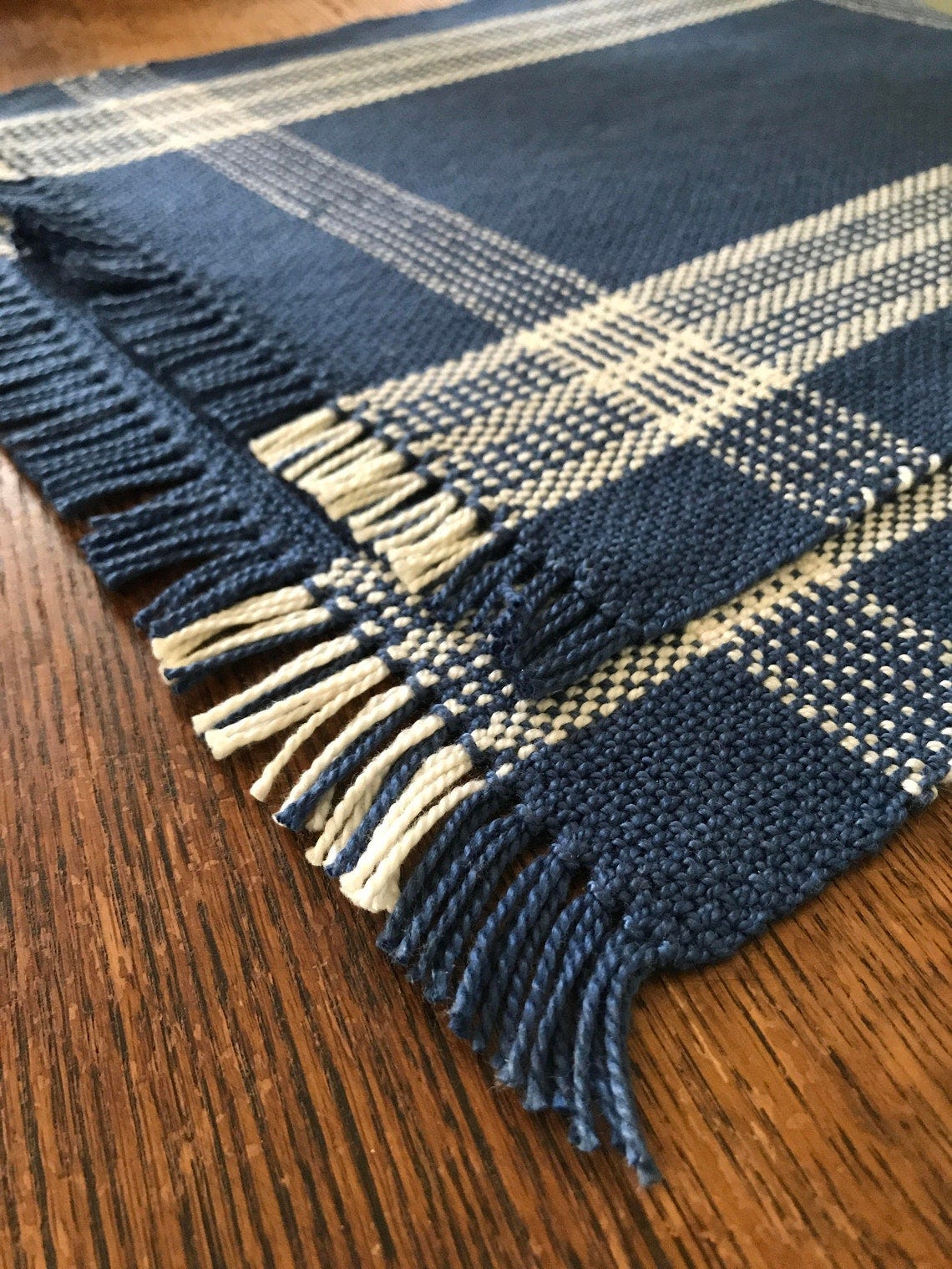 May include: A blue and white plaid placemat with fringe edges. The placemat is woven with a simple pattern and has a slightly textured surface.