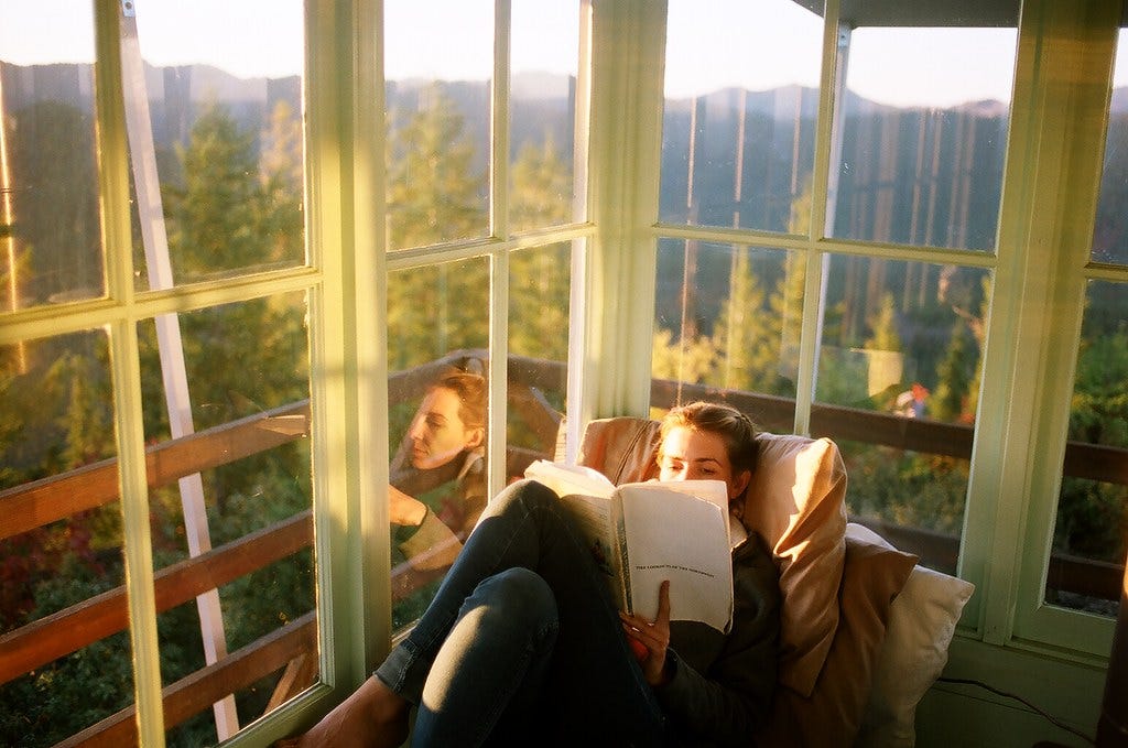 A woman reading a book while relaxing on cushions in a sun-filled window nook with a scenic nature view outside.