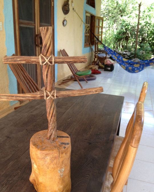 A cross made of twisted vine with two crossbars lashed with twine, set in a log, on a dark wood table on a tiled porch, a blue hammock behind. A cross made of twisted vine with two crossbars lashed with twine, set in a log, on a dark wood table on a tiled porch, a blue hammock behind.