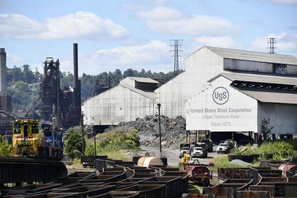 Lines of empty carts on a railroad with three white U.S. Steel buildings in the background, one with blue writing that says "United States Steel Corporation" on the side.