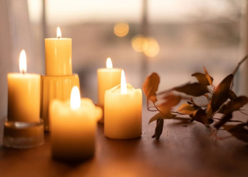 Lit candles on a wooden table with soft bokeh background. Warm glow creates a cozy, tranquil atmosphere. Nearby are dried leaves.