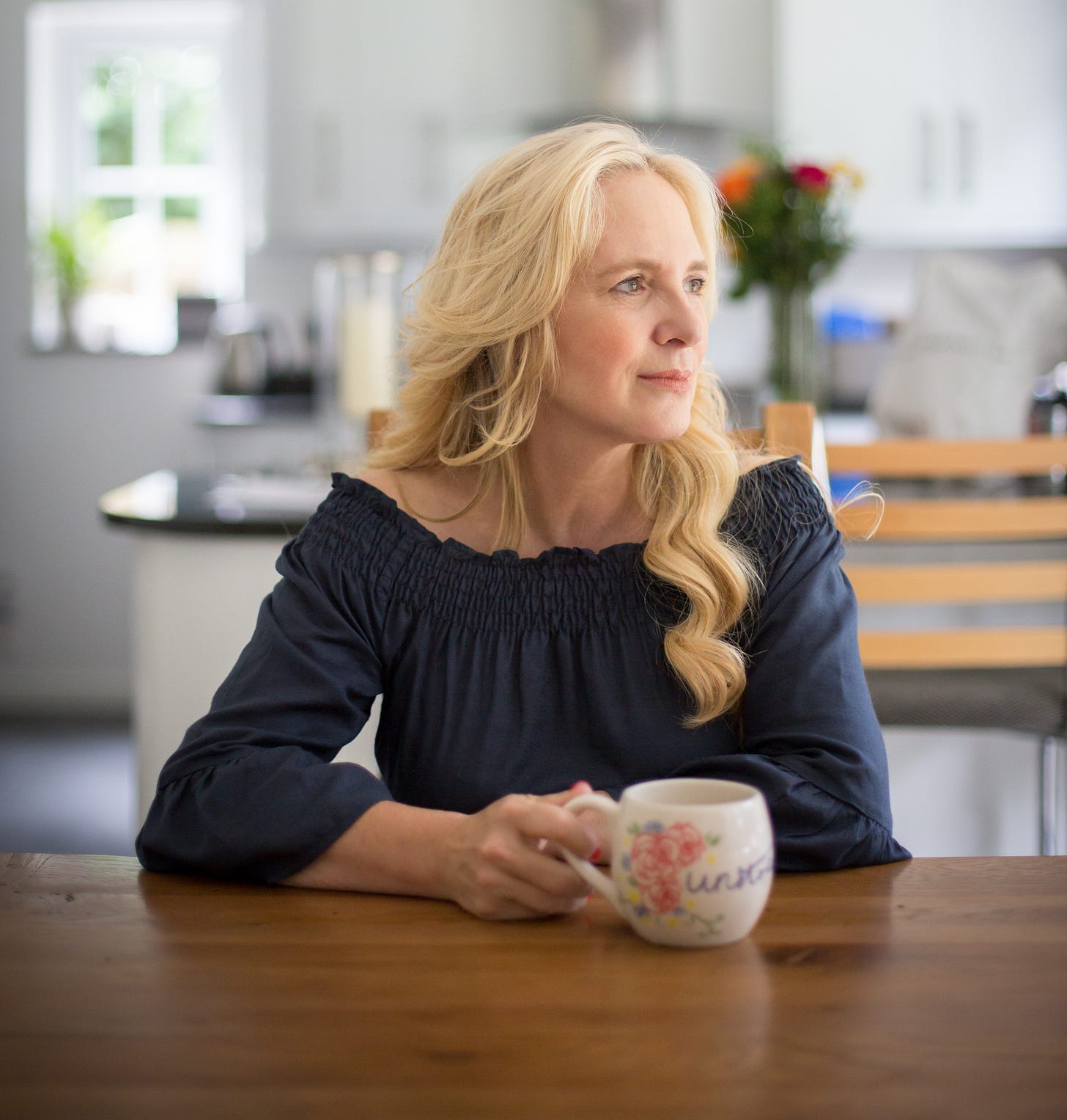 Danusia Malina-Derben sitting at a wooden table with a mug, in a light-filled kitchen, looking into the distance