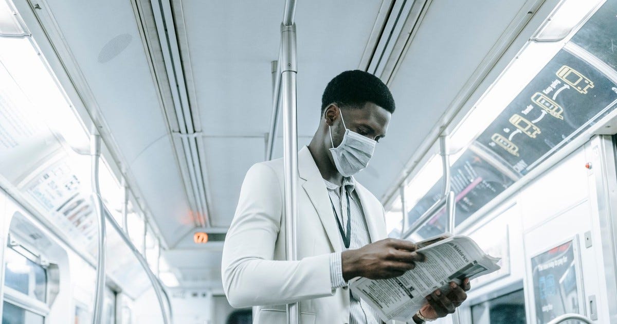 African American man in a mask reading a newspaper on a train during the COVID-19 pandemic.