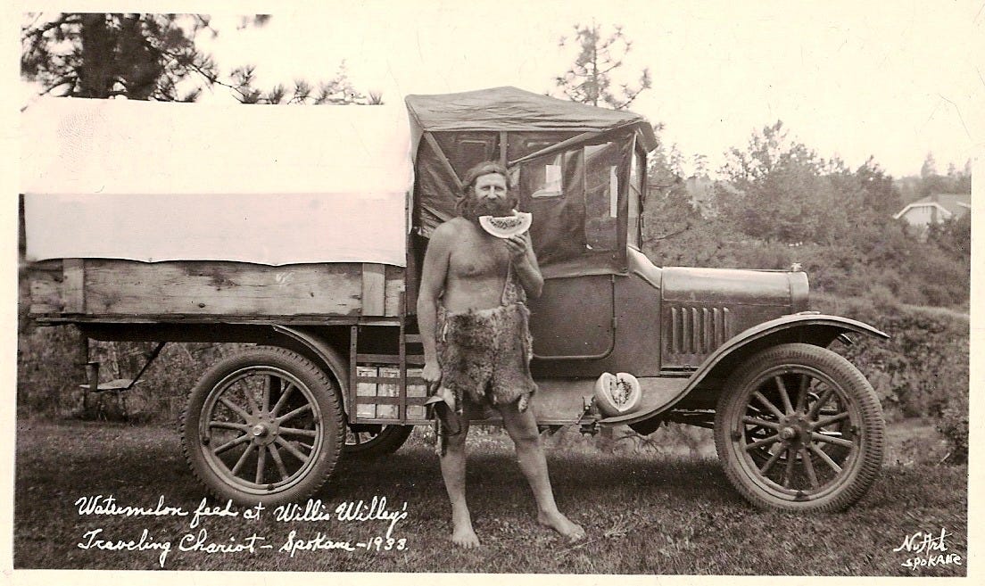Black-and-white postcard image of Willie Willey standing barefoot and bare-chested beside his modified truck, wearing a rough animal-hide wrap and holding a slice of watermelon. The vehicle behind him is outfitted as a traveling home, with handwritten text identifying the scene as “Watermelon feed at Willie Willey’s Traveling Chariot” in Spokane, 1933.