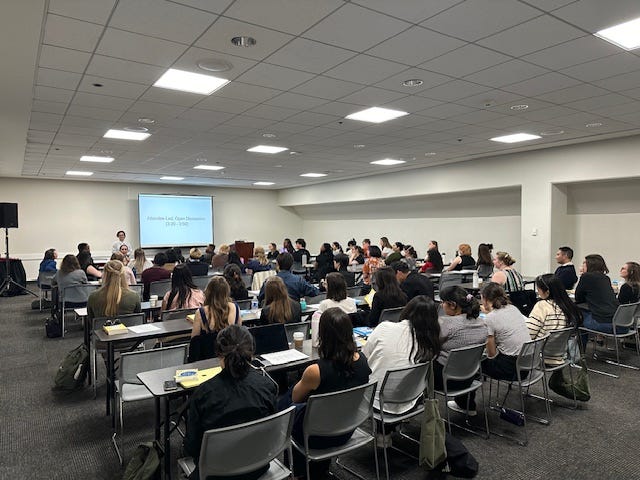 Conference room full of seated people at long tables facing a single speaker at the front of the room.