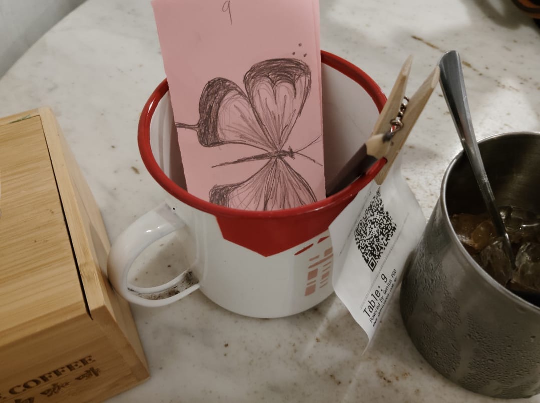 A sketch of a butterfly on a pink piece of paper meant for writing one's order on in a cup. A coffee mug and a tissue box with 'coffee' written on it are visible to the side.