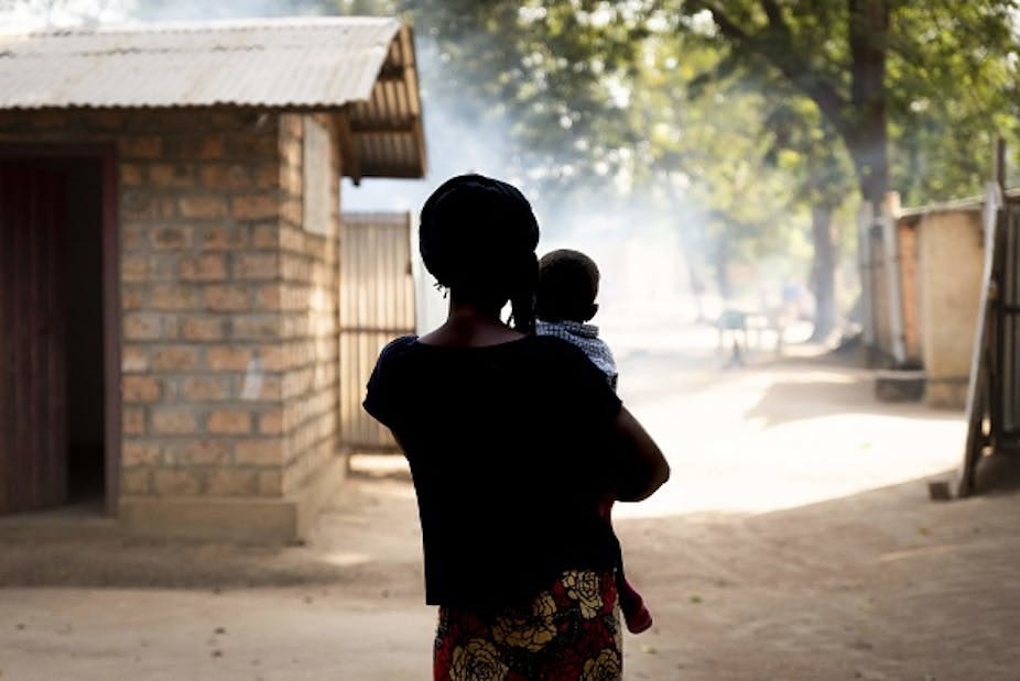 View from behind of a woman holding a baby, standing in an unpaved space between simple buildings. View from behind of a woman holding a baby, standing in an unpaved space between simple buildings.