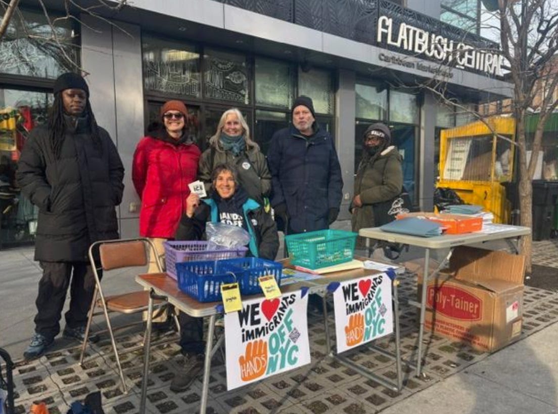 Image of men and women in front of a table with two signs that say " says "We heart immigrants. Hands off NYC" with a hand on it" On the table are baskets with whistles and zines. Image of men and women in front of a table with two signs that say " says "We heart immigrants. Hands off NYC" with a hand on it" On the table are baskets with whistles and zines.