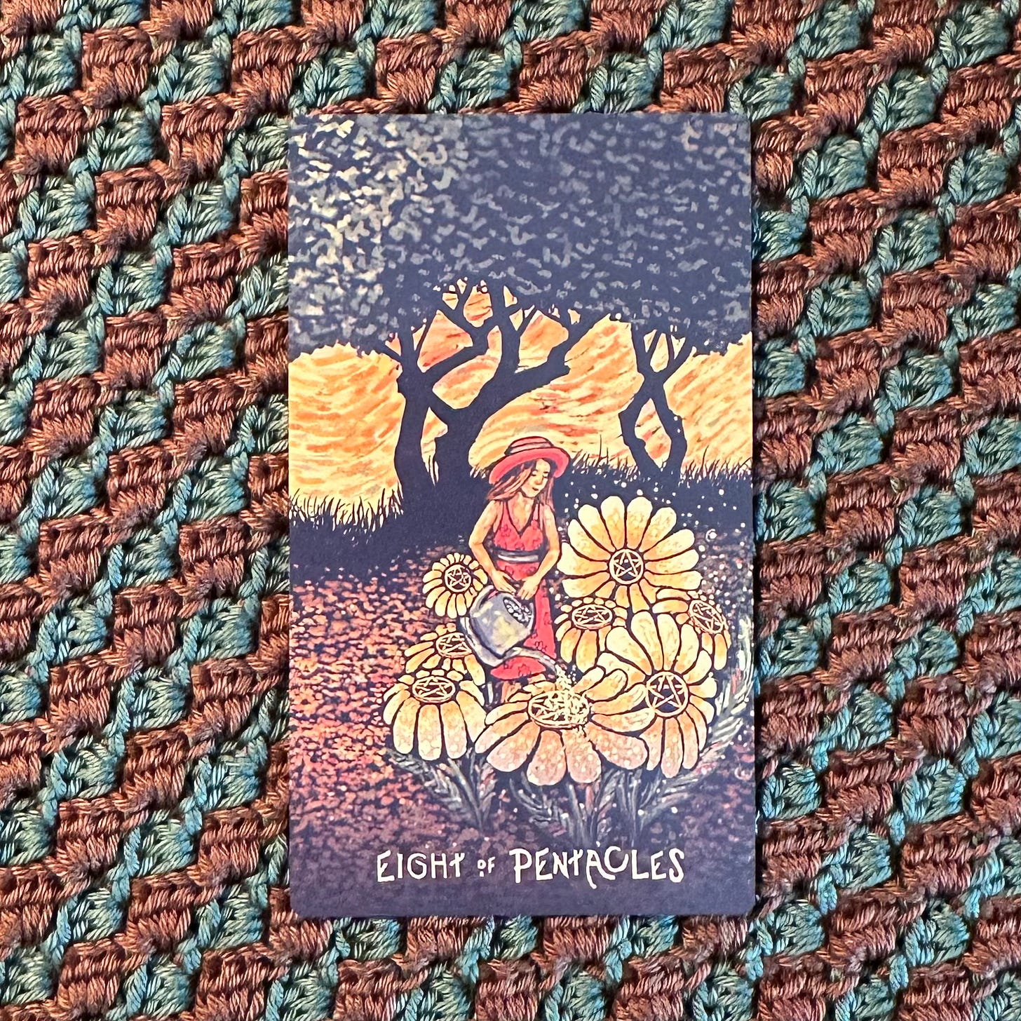 A young woman in a red sun hat and dress stands in a field, in front of some trees and golden sky, watering eight full open golden flowers, each with a pentacle at their centre