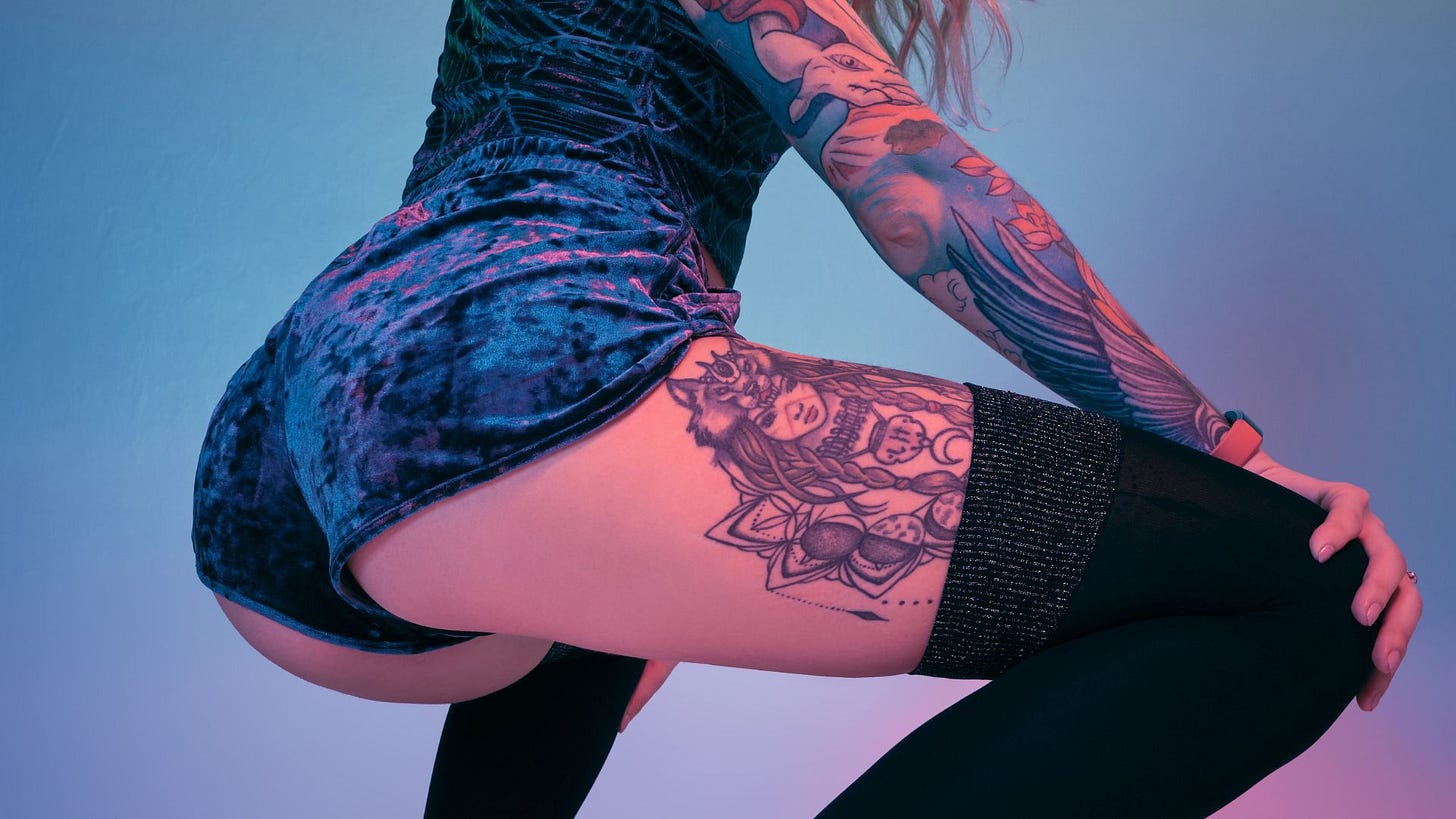 Light blue background, photo from behind of a fine, round butt in the twerk position, dark blue vervet short shorts, black leggins, and classy ink on right arm shows blue wings, on the thigh is of a Goddess.
