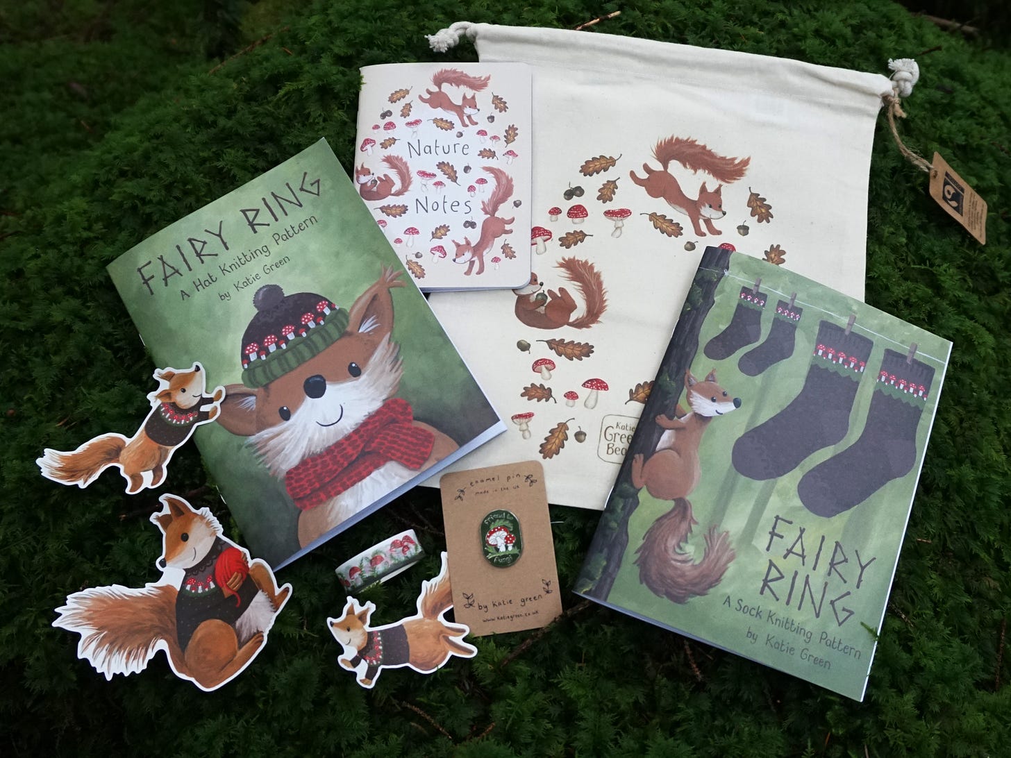 Image description: A pile of cute goods laid out on a bed of squishy green moss. There is an unbleached cotton drawstring project bag printed with tumbling squirrels, leaves and mushrooms. There's a little notebook entitled Nature Notes with the same squirrl illustration on the front. There are two illustrated pattern booklets, three cute illustrated stickers of squirrels wearing cardigans, a roll of mushroom washi tape and an enamel pin.