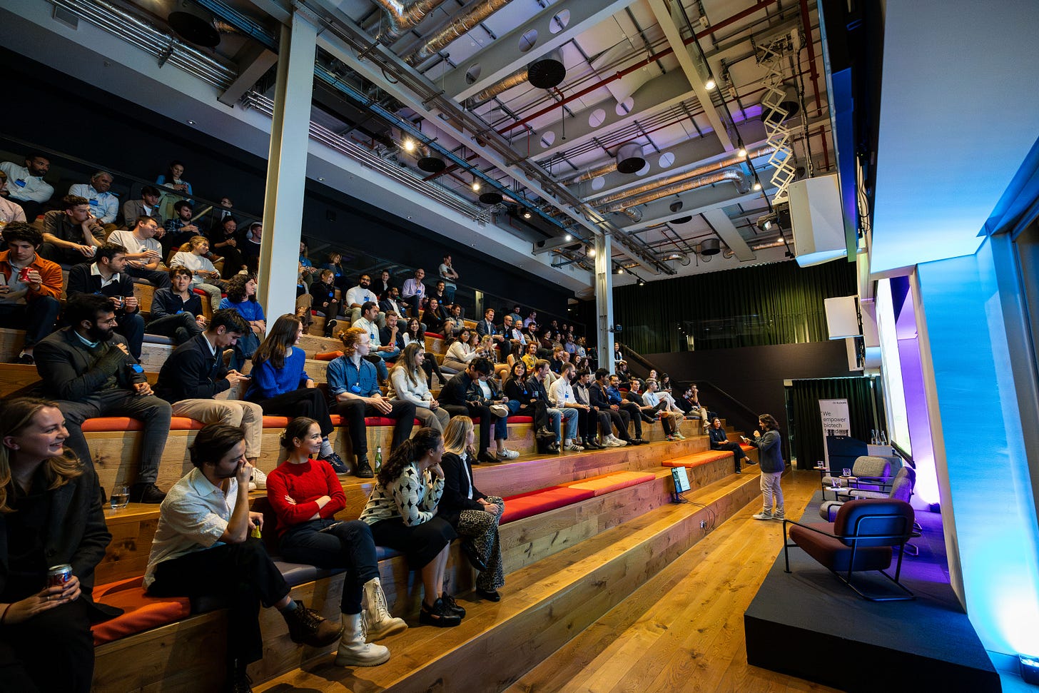 A photo of an audience sat on staggered seating and listening to a person standing at the front. 