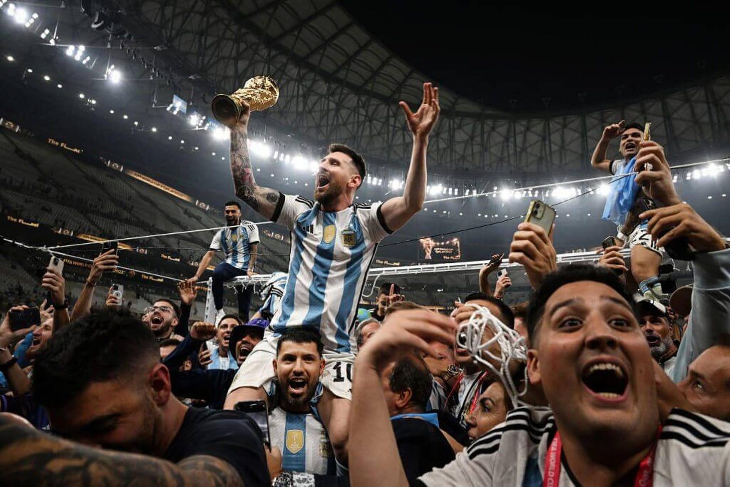 Lionel Messi celebrates with Argentina fans and team-mates, lifting the trophy in his right hand under the spotlights in the Lusail Stadium on Sergio Ageuro's shoulders after winning the World Cup in 2022