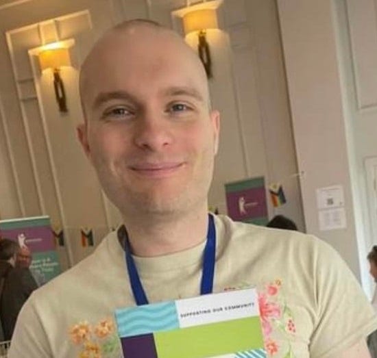 A photo of Scott Neilson. They are smiling and holding a copy of their Creating Safe Spaces for Autistic People book.