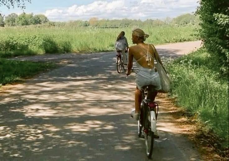 This may contain: two people riding bikes down a dirt road next to tall grass and green trees in the background