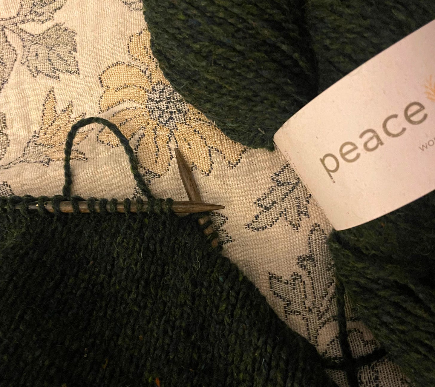 A close up image of a knitting project in dark green wool on a floral blanket. There is an unwound hank of the dark yarn with the label still on, that reads Peace Fleece.