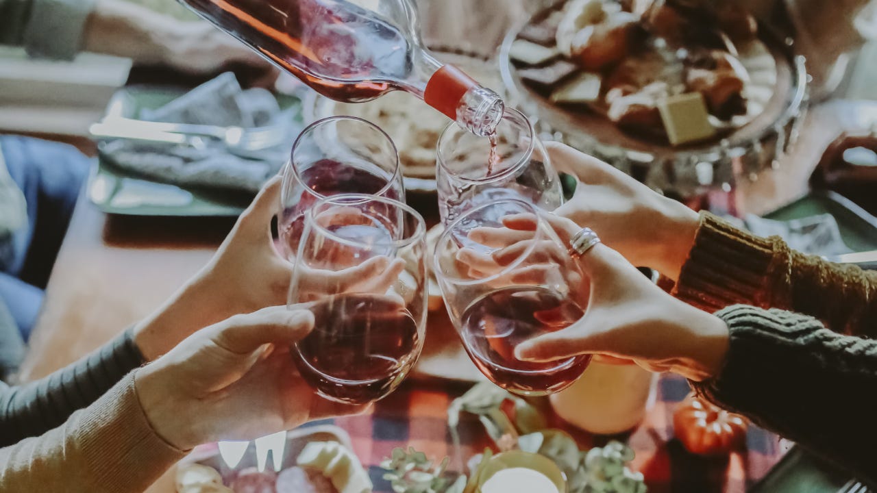 **Alt text:** People raising glasses of red wine around a dinner table while a bottle pours more wine during a shared meal.
