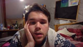 A lightly bearded, tired young man is laying on his bed facing the camera.
