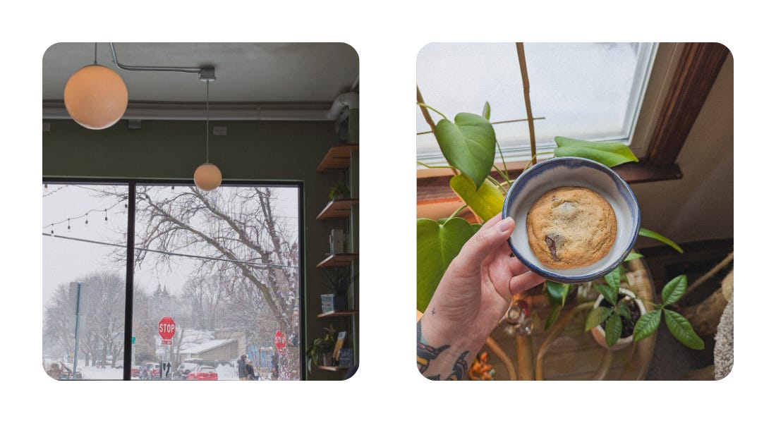 Image 1: the inside of a cafe with two orb lights hanging from the ceiling, visible in the back is the large picture window where it is snowing outside and trees and stop signs are visible. Image 2: Alej's hand holding a blue and white bowl with a chocolate chip cookie, plants are in the background, their arm is tattooed