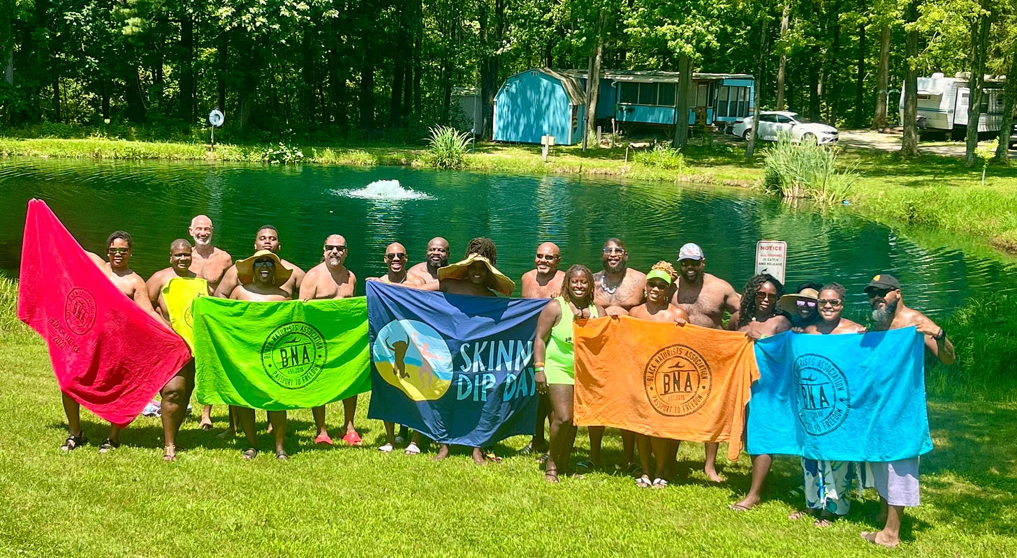 A large group of nude and partially draped people stand smiling on a grassy lawn beside a wooded pond, holding up colorful towels. Several towels are emblazoned with the Black Naturists Association logo in red, green, orange, and blue, and a central navy towel reads "Skinny Dip Day." A fountain sprays in the pond behind them. A large group of nude and partially draped people stand smiling on a grassy lawn beside a wooded pond, holding up colorful towels. Several towels are emblazoned with the Black Naturists Association logo in red, green, orange, and blue, and a central navy towel reads "Skinny Dip Day." A fountain sprays in the pond behind them.