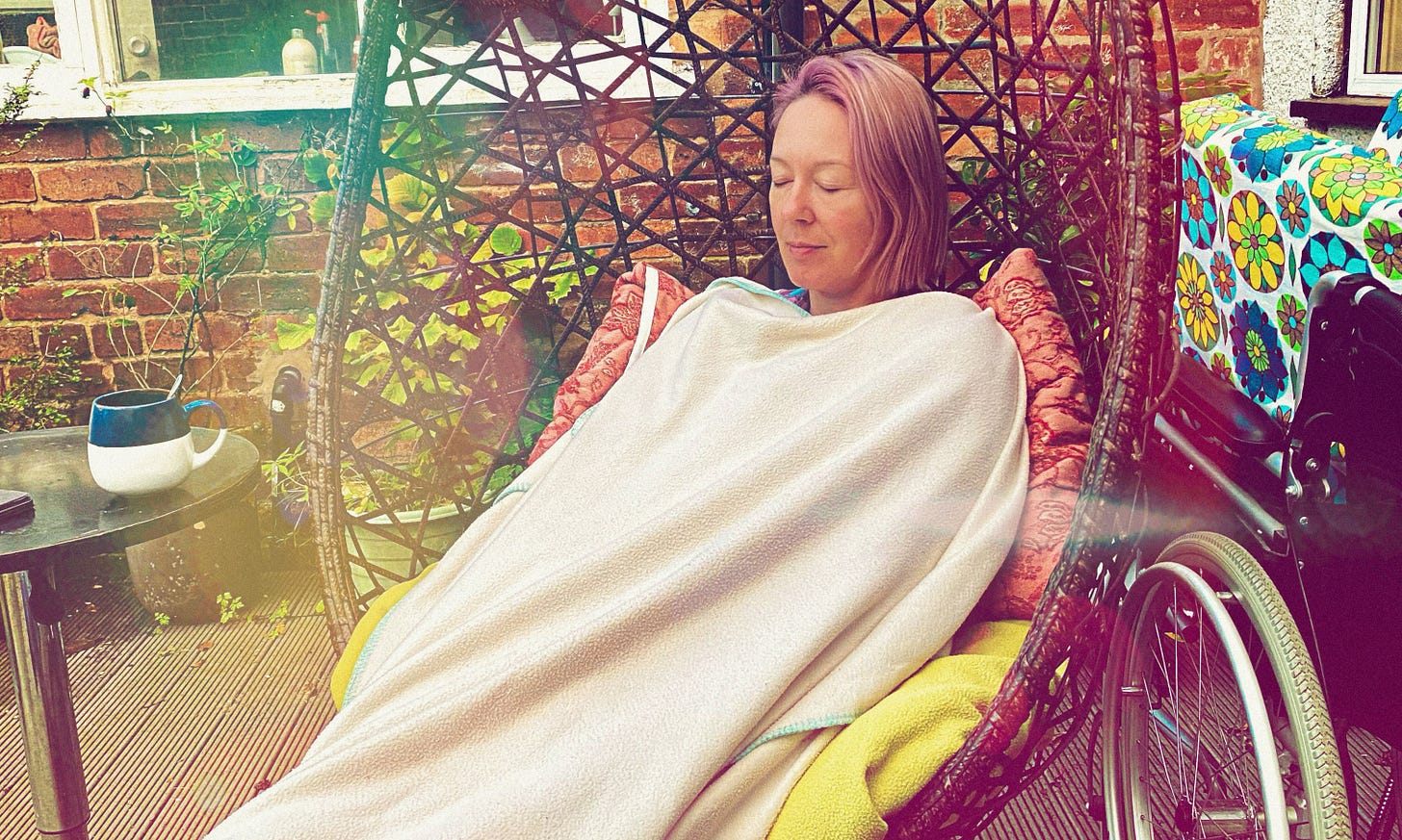 hotograph of Yvie, a white female with shoulder-length pink hair is sitting against pillows in a large egg chair covered in a large white blanket. Her wheelchair is on her left. A table with a mug is on her right. Garden decking setting. Her eyes are closed. She is resting. hotograph of Yvie, a white female with shoulder-length pink hair is sitting against pillows in a large egg chair covered in a large white blanket. Her wheelchair is on her left. A table with a mug is on her right. Garden decking setting. Her eyes are closed. She is resting.
