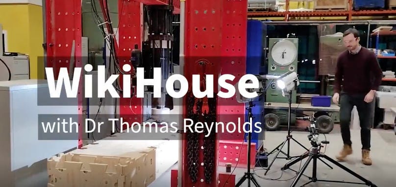 Still from a video, it reads WikiHouse with Dr Thomas Reynolds - Thomas can be seen in the background standing in a lab. He is about to test a WikiHouse block