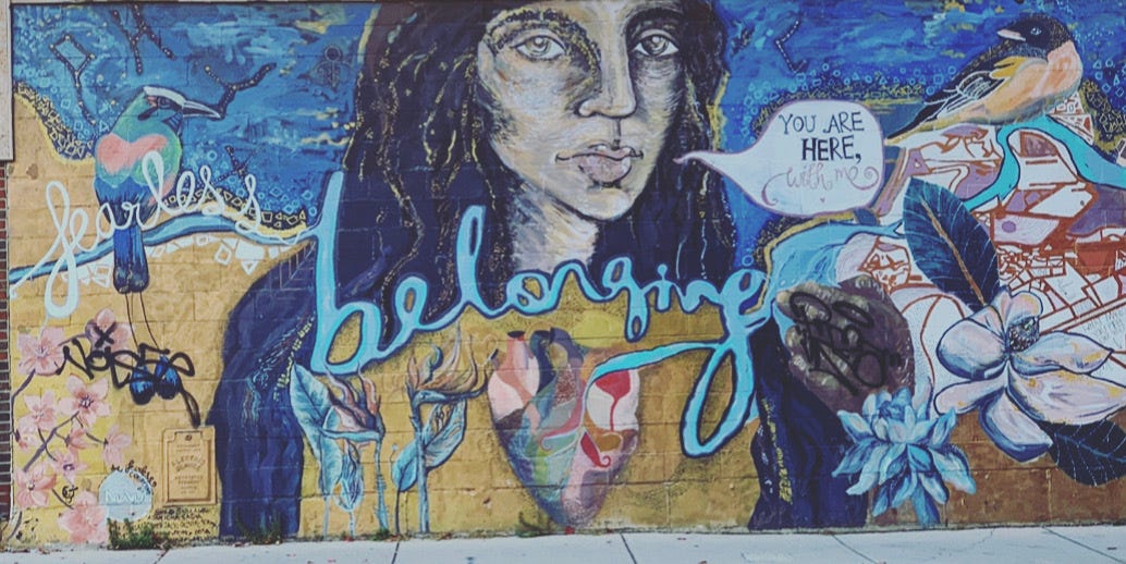 A mural of a person with long dark hair and a multicolored hear that says "belonging" across their chest. They are surrounded by flowers and stars and birds