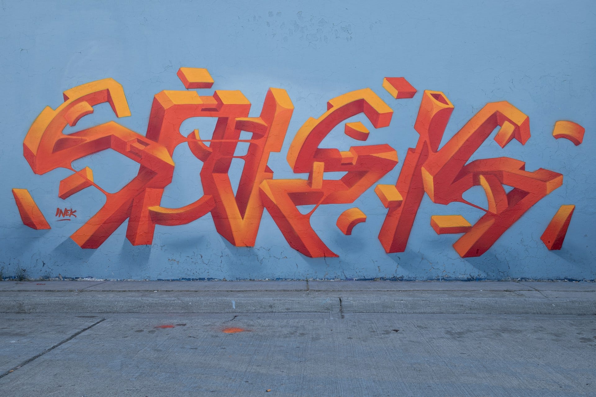 Un’opera di street art in Messico, firmata da Inek. Su uno sfondo celeste si staglia l’illustrazione di forme rosse in 3D che assomigliano a delle lettere astratte. Un’opera di street art in Messico, firmata da Inek. Su uno sfondo celeste si staglia l’illustrazione di forme rosse in 3D che assomigliano a delle lettere astratte.