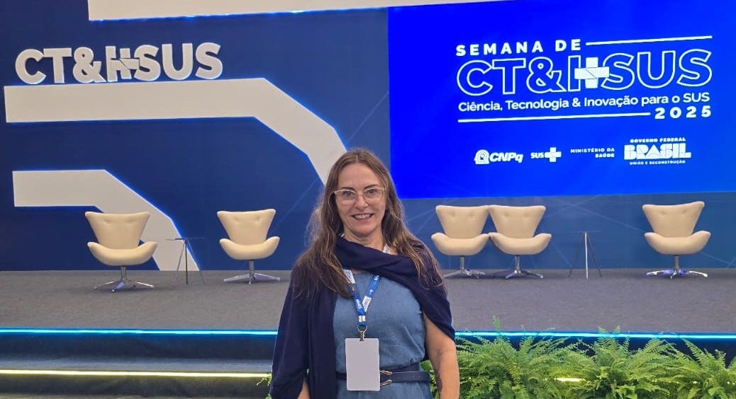 Descrição da imagem: Cristina Rabelais é uma mulher branca, de cabelos castanhos e longos. Usa óculos de grau, um vestido jeans e uma echarpe azul-marinho. Ao fundo, há o palco do evento com um painel azul e branco e um telão exibindo a logo da Semana de CT&I para o SUS 2025 e seis poltronas brancas. Na base do palco, um conjunto de samambaias.
