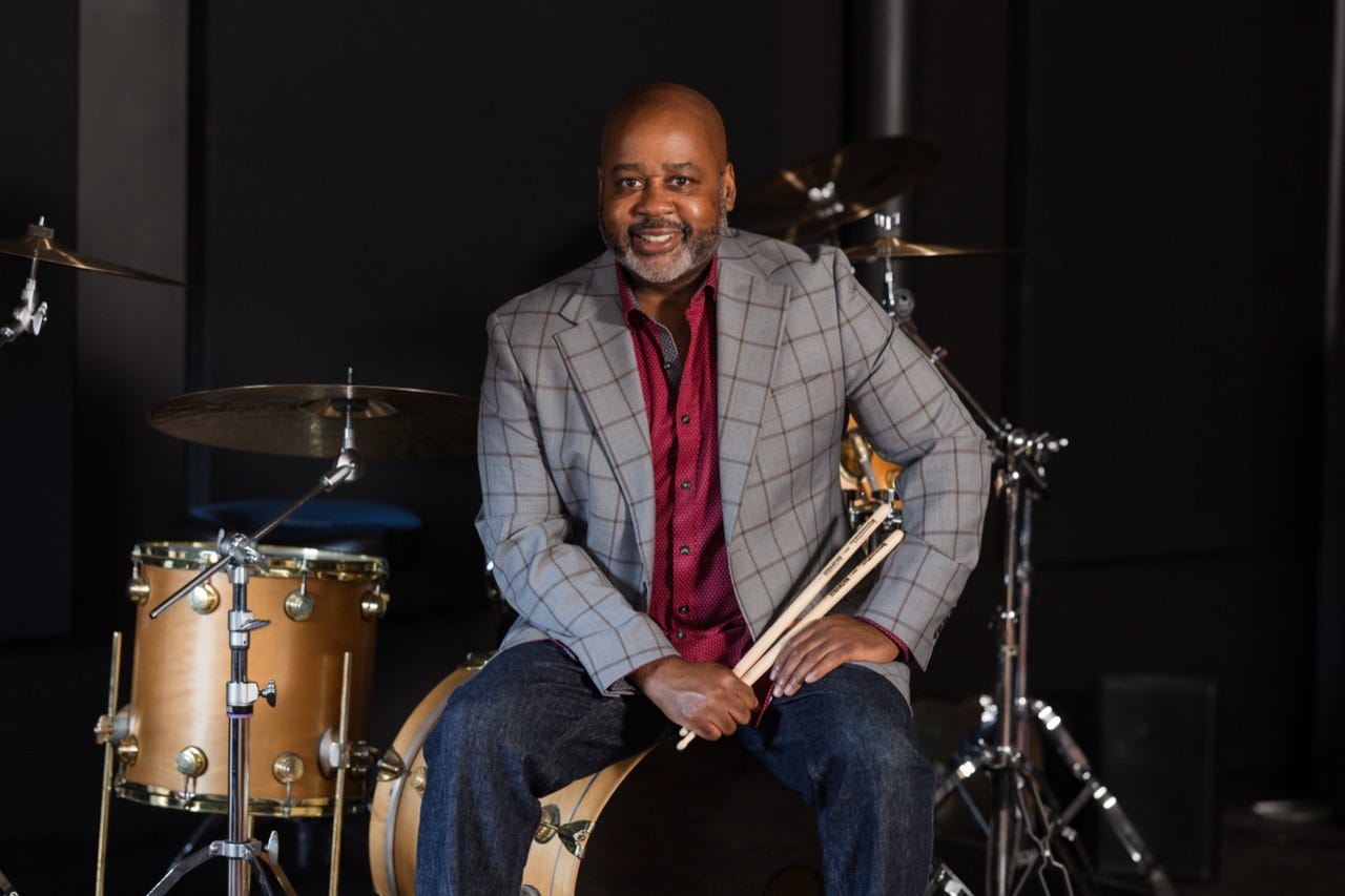 Clayton Craddock smiling, seated at his drum kit in a gray plaid jacket and red shirt Clayton Craddock smiling, seated at his drum kit in a gray plaid jacket and red shirt
