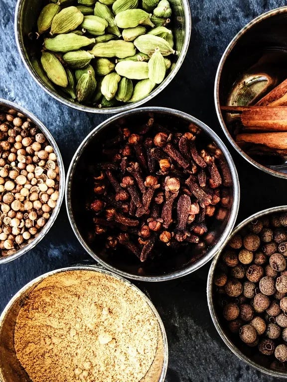Arial picture, close up of small steel pots holding the spices that together make mixed spice. Clockwise from the top: green cardamom pods, cinnamon quills, clove buds, allspice berries, ground ginger, coriander seeds