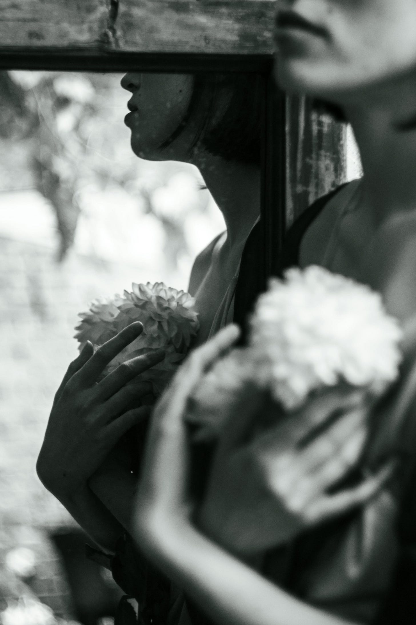 A blurred black-and-white reflection of a woman holding soft white flowers to her chest, partially obscured by a mirror—evoking themes of identity, disappearance, and gentle reclamation.