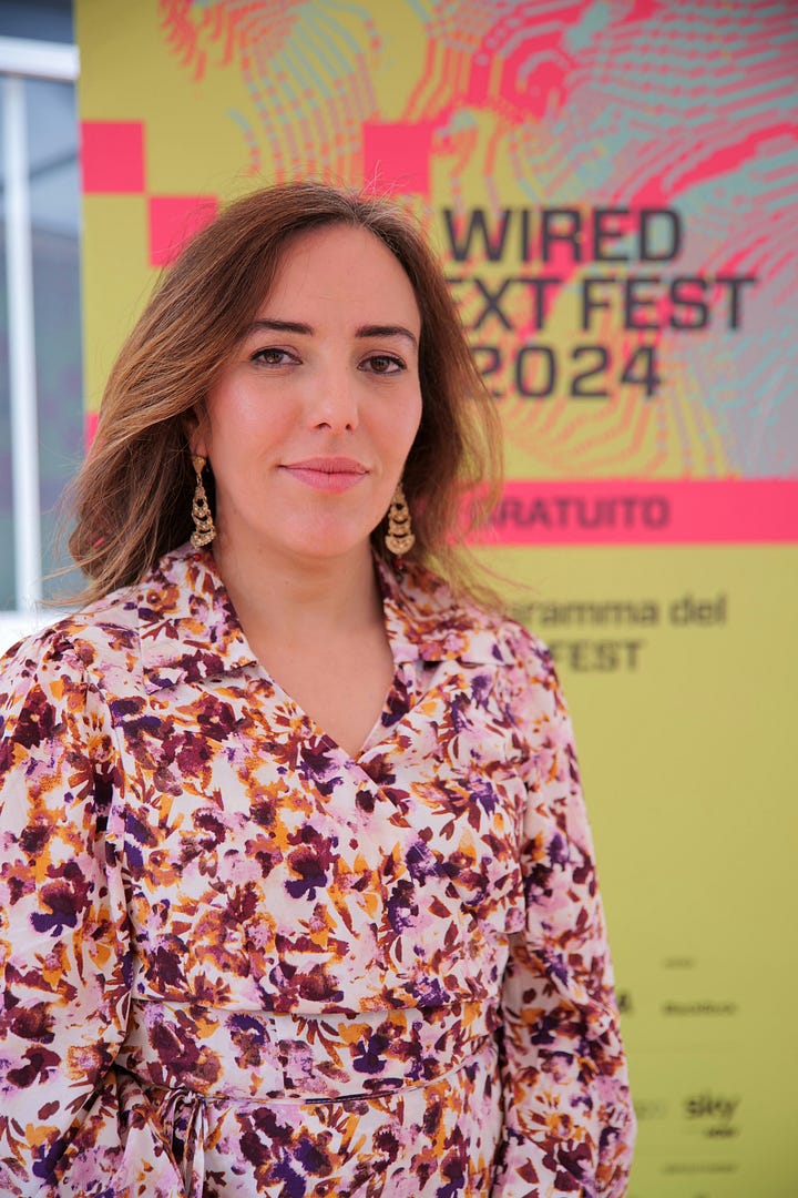 Stella Assange next to a brick wall. Stella Assange in front of a yellow Anything to Say banner holding a Free Assange black tote. Stella Assange in front of a Wired Italia 2024 poster. Stella Assange smiling leaning against a collumn.