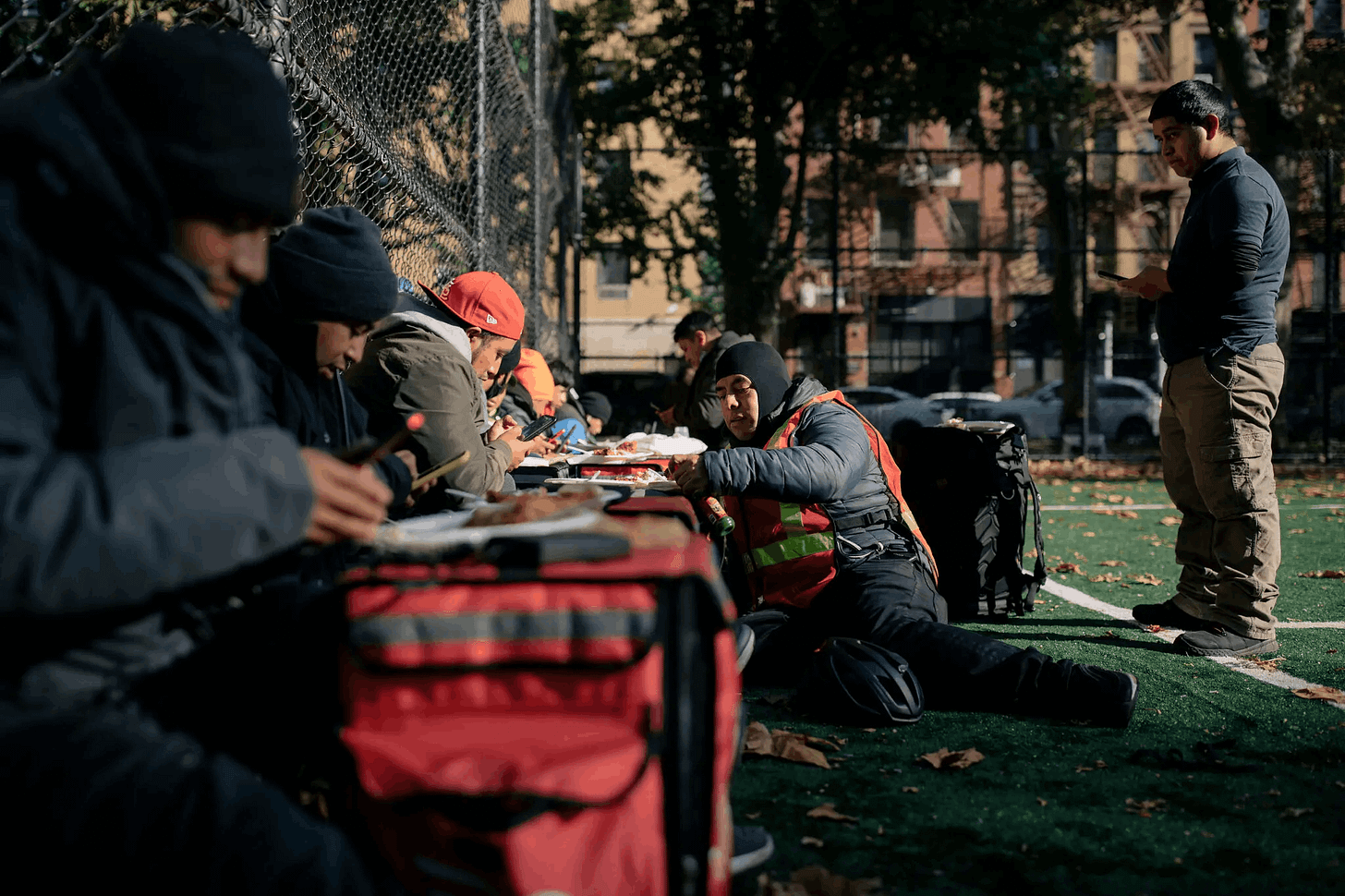 Image depicts a line of delivery drivers sitting on a park ground and eating as they wait for their next orders. Image depicts a line of delivery drivers sitting on a park ground and eating as they wait for their next orders.
