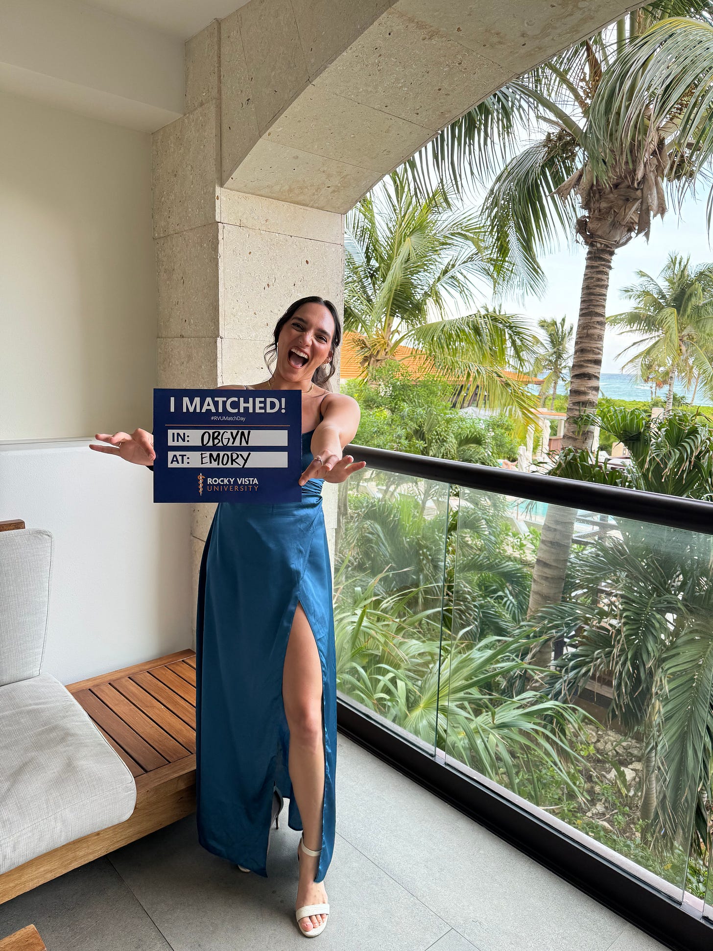 Em in a blue dress on a balcony smiling and holding a sign that says I match in OBGYN at EMORY