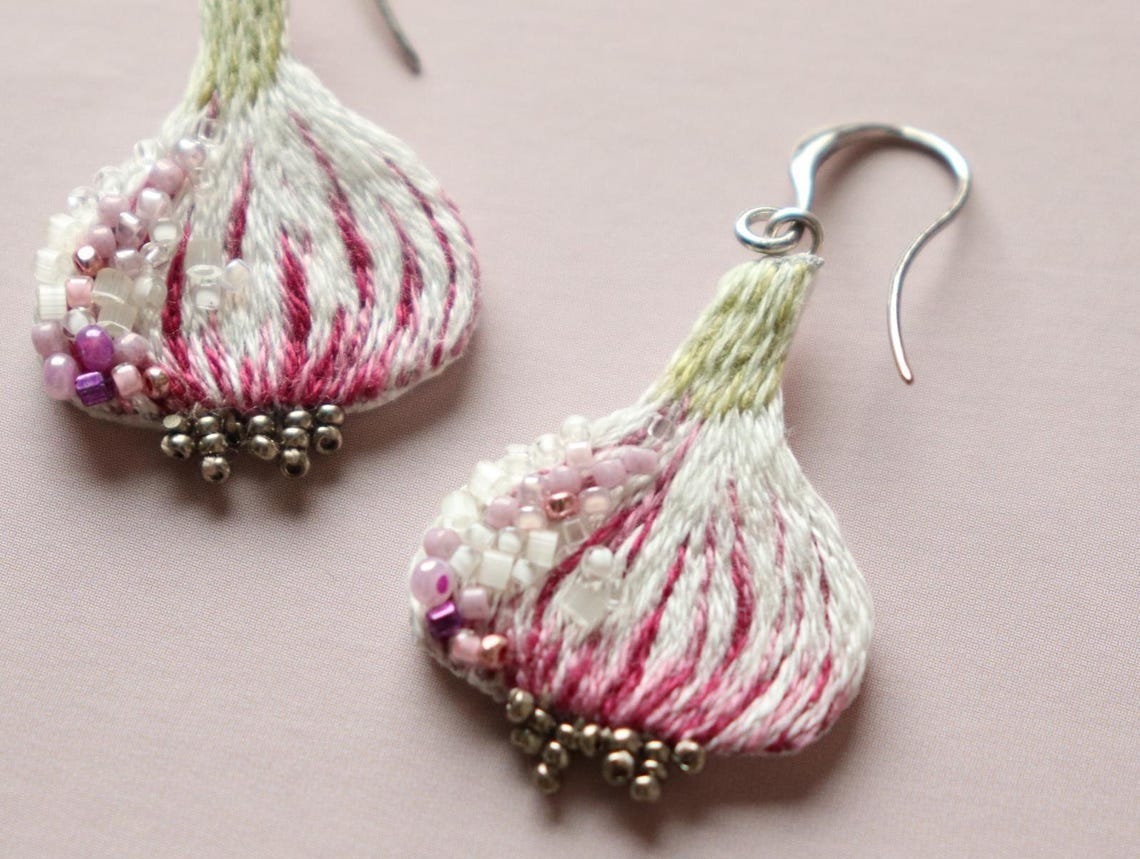 May include: A pair of pink and white floral earrings with silver hooks. The earrings are made of embroidered fabric and beads.