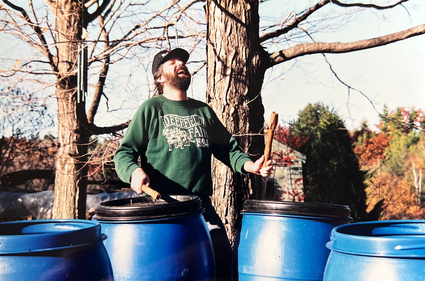 Uncle Kevin outside using sticks to bang heartily with great laughter on a bunch of large blue barrels with lids