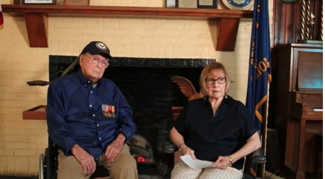 An elderly man and woman are seated indoors in front of a black fireplace with a yellow brick wall. The man is wearing a navy blue shirt, khaki pants, a dark cap with an emblem, and has military medals pinned to his chest. The woman is wearing glasses, a dark blouse, and light-striped pants, and is holding papers. There are plaques and flags displayed in the background, as well as a piano to the right. The scene has a formal yet relaxed atmosphere, possibly indicating a commemorative or interview setting. No specific recognizable figures, landmarks, or brands are visible.